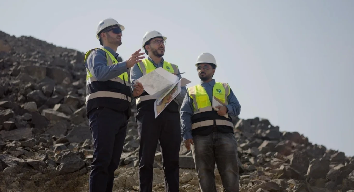 ثلاثة مهندسين يرتدون سترات عاكسة وخوذات بيضاء يقفون على أرض صخرية، أحدهم يشير والآخر يحمل خرائط.