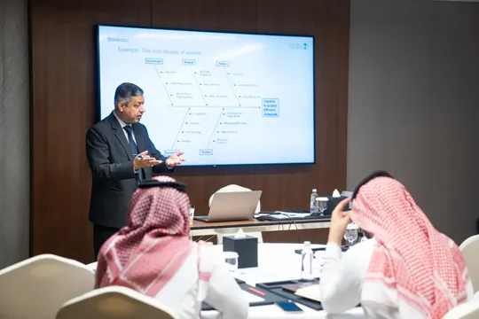 عرض بيئي توضيحي رجل يرتدي بدلة يقدم عرضًا تقديميًا أمام شاشة كبيرة تعرض مخططًا بيانيًا، بينما يستمع إليه ثلاثة رجال يرتدون الزي السعودي.
