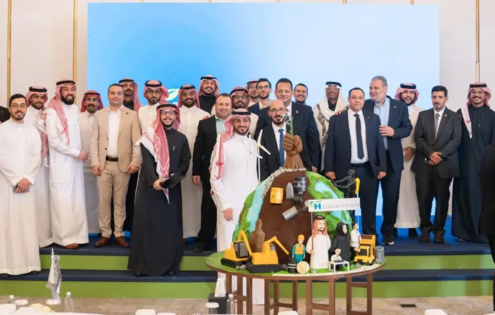 مجموعة من الرجال السعوديين يرتدون الزي التقليدي والبدلات، يقفون حول كعكة كبيرة على شكل كرة أرضية مزينة برموز بيئية وصناعية.